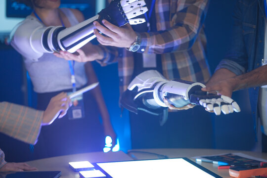 An Indian woman wearing a robotic arm prototype shakes hands with another robotic arm held by a colleague. A diverse tech team smiles, collaborating in a lab.