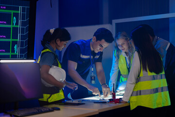 A diverse team of engineers in safety vests collaborates at night. They are pointing at a blueprint...