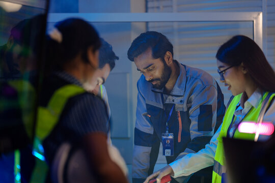 An Indian supervisor leads a night shift briefing in a smart factory control room. He discusses an AI automation system or incident response with his female engineers.