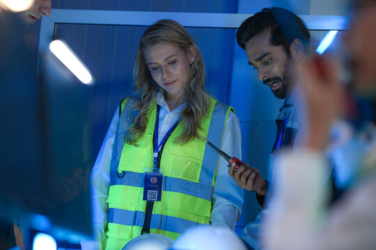An Indian engineer with a radio points at a light table, reviewing a blueprint with his smiling Caucasian colleague in a smart factory control room.