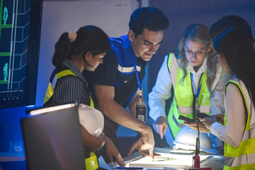 A large logistics team reviews supply chain blueprints. The diverse group, including an Indian...