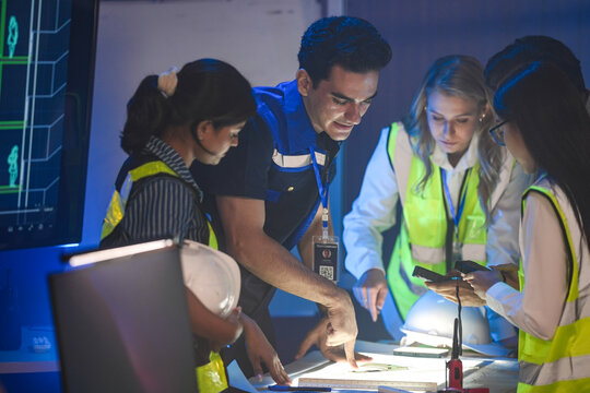 A large logistics team reviews supply chain blueprints. The diverse group, including an Indian woman with a hardhat, plans a new AI strategy for smart factory optimization.