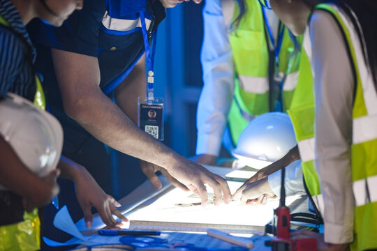 A close up of engineers reviewing a CAD blueprint on a light table. The team is in a smart factory SOC, analyzing an AI automation system and using a radio.