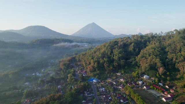 Stunning aerial drone footage of Bajawa village in Flores, Indonesia. A misty valley and the iconic Mount Inerie volcano are visible in the background during a beautiful golden hour sunrise.
