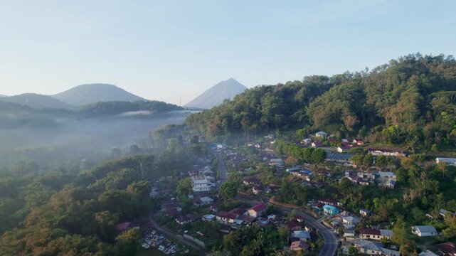 Aerial drone footage ascends, revealing the iconic Mount Inerie volcano from behind a hill. The village of Bajawa, Flores, is visible below in a misty valley at sunrise in Indonesia.
