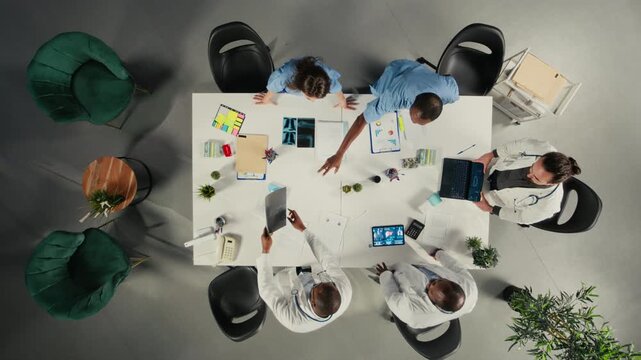 Top down view of medical experts group review clinical data, working together and sharing their expertise to improve healthcare services at the hospital. Brainstorm treatment ideas. Camera A.