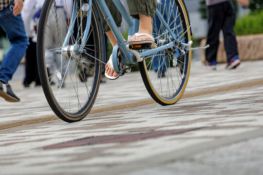 Ground level view of flip flop wearing cyclist pedaling a light blue bike across a tiled patterned city walkway bustling with pedestrians passing nearby in bokeh background. - Powered by Adobe