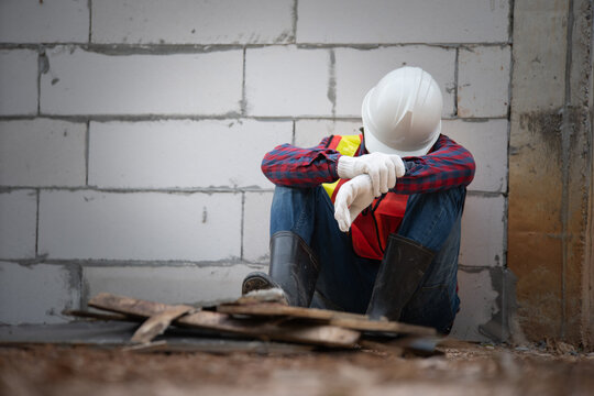 A construction worker sits against a wall, wearing a hard hat and safety gear