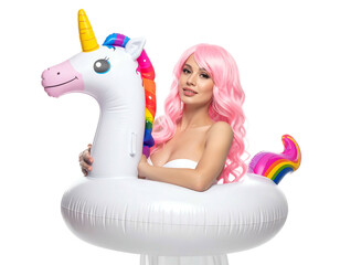 Woman with pink curly wig, white fabric wrap, holds inflatable unicorn float, bright studio lighting, black background