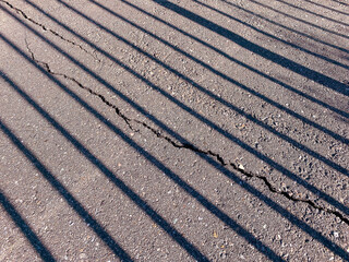 Shadows from vertical bars create striped patterns across a cracked asphalt surface sunlight casts dark lines while the rough pavement texture and crack add visual interest
