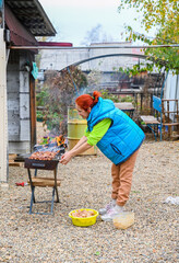A 50-year-old Russian woman in a rural village grills marinated chicken outdoors, tending skewers over an open flame on a cool autumn day in Russia.