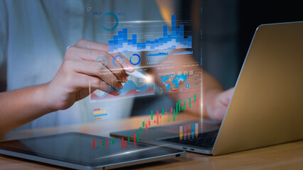 Business analyst using digital tablet and laptop with holographic data charts and financial analytics dashboard, representing big data visualization, fintech investment, and smart business strategy.
