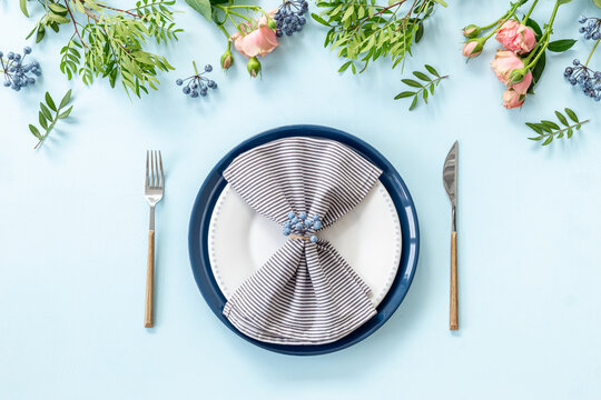 Top view of table setting with empty plate, cutlery and napkin - pink roses flowers decorated on blue background, flat lay