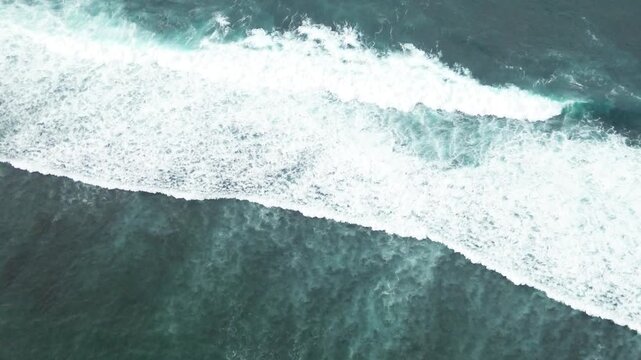 Aerial view of ocean waves crashing in Uluwatu, Bali. The turquoise water and white foam create a stunning natural pattern from above.