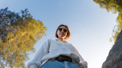 Low angle wide shot of young woman wearing sunglasses and casual outfit standing outdoors, surrounded by tall green trees and clear blue sky, confident and relaxed mood
