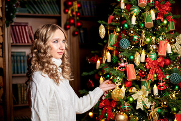 girl in a white sweater is sitting in an armchair next to a Christmas tree. The Christmas tree is decorated with white and red balloons and gifts. The girl has a Christmas present in her hands.