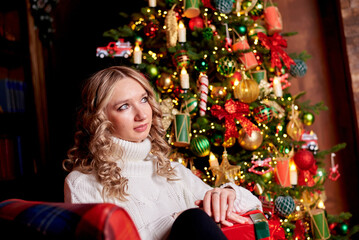 girl in a white sweater is sitting in an armchair next to a Christmas tree. The Christmas tree is...