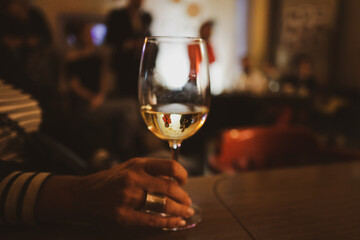 Shallow depth of field (selective focus) details with a glass of white in the hands of a woman at a party with people visible in glass.