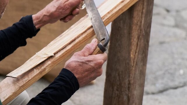 Bottom-to-top pan following an elderly artisan shaving a log with a drawknife to create wood splints for basket weaving; curling chips, textured hands, outdoor daylight.