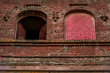 Ein dramatisches Nahaufnahme-Detail einer alten Backsteinmauer aus dunkelrotem Ziegelwerk, das von Verwitterung und rötlichem Putz überzogen ist. Die Struktur zeigt zwei Bögen: Der linke Bogen öffnet 