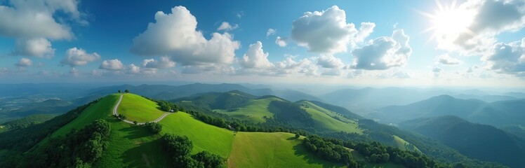 Fototapeta premium Aerial panorama shows rich green rolling hills under bright blue sky. White clouds drift over distant hazy mountains. A winding path leads to hill peak. Sun shines over peaceful vast landscape.