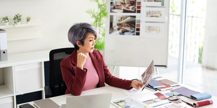 Smiling asian woman interior designer sit at modern office desk considering document in morning. Happy female architect work with paperwork use laptop working on a project at workplace. - Powered by Adobe