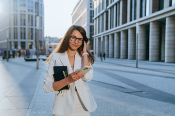 A businesswoman stands outdoors, holding a smartphone and notebook, dressed in a white suit, embodying elegance and modern professional life.