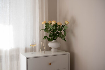 Vase with champagne roses and glass of white wine, candles on dresser table in bedroom. Interior element