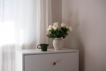 Vase with white roses with cup of coffee on dresser table in bedroom. Interior element