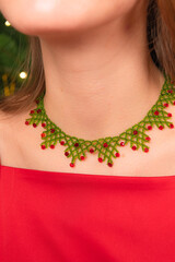 Close up on a neck of a girl in red dress wearing red and green beaded necklace like a spider web next to the Christmas tree, ethnic handmade concept.