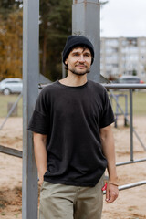 Casual bearded man in black t-shirt and beanie standing confidently in outdoor urban park setting, autumn background, relaxed streetwear look 