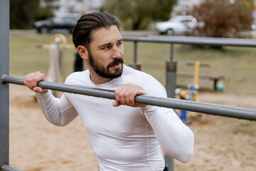 Close-up of a young bearded man in a white compression shirt doing pull-up exercise on outdoor fitness bars, focusing on upper body strength and determination in a park setting
