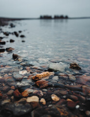 Pebble-strewn lakeshore under overcast sky with distant cityscape and calm water, capturing natural textures and tranquil outdoor atmosphere