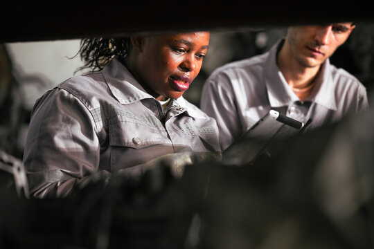 Female technician check used car damaged engine block at scrapyard warehouse recycle area part. African American engineer inspecting rusty oily auto motor old spare part in junkyard for reuse service