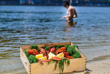
A box of boiled crayfish with herbs and lemons on the riverbank, a girl swimming in the background