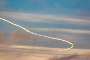 A dramatic jet contrail arcs across a serene, colorful sky with wispy clouds, creating a dynamic visual against the peaceful backdrop
