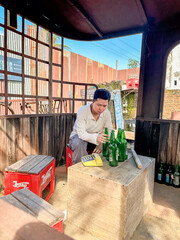 A young man is sorting green glass bottles on a wooden table in a casual outdoor space