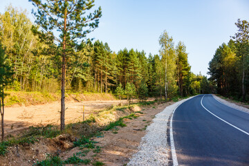 Expansive plot cleared for development by a winding road. Lush forest fringes the construction site under a bright summer sky