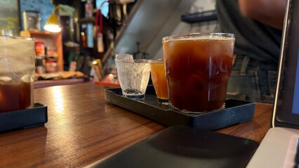 A flight of cold beverages, including iced coffee, served on a tray on a wooden table, suggesting a tasting session or break while working on a laptop in a cozy cafe.