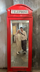 A couple stands together, taking a selfie in a bright red vintage phone booth with a mirror