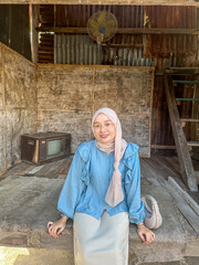 A woman in a blue blouse and hijab sits on a wooden floor, smiling in a quaint, rustic setting