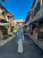 A young woman in a hijab walks down a narrow street of wooden homes, basking in the sun