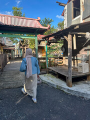 A woman in a hijab strolls through a rustic village, surrounded by wooden buildings and greenery