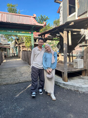 A young couple stands smiling in a traditional village, surrounded by wooden houses and greenery