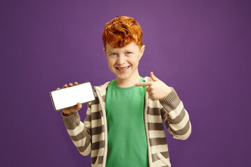 Isolated Portrait of Joyful Red Haired Child Boy Holding Smartphone and Showing to Camera Empty Screen of it Display, Points There With her Finger, Smiles Standing Against Violet Isolated Background