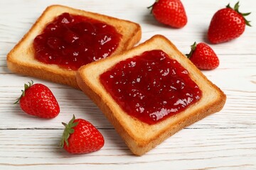 Two slices of toasted bread with strawberry jam and fresh whole strawberries on a white wooden table