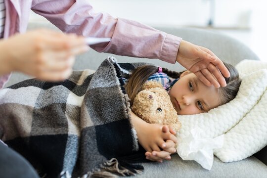 A young girl lies on a couch, wrapped in a blanket, while her mother checks her forehead for a fever. A teddy bear is held close.