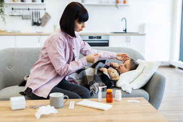 A concerned mother checks her daughter's temperature while the child rests on a couch, surrounded by medicine and tissues.
