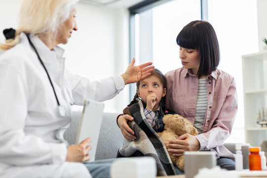 A concerned mother holds her sick daughter while a doctor checks her temperature. The child is wrapped in a blanket.