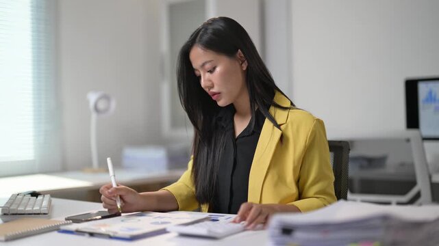 A woman is sitting at a desk and writing with a pen. She is wearing a yellow jacket and she is focused on her work. The desk is cluttered with papers and a calculator